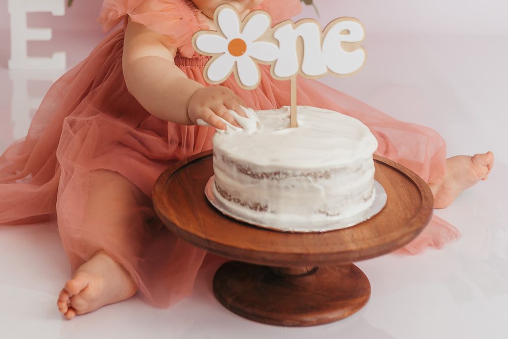 Baby girl in pink tulle dress touching her first birthday smash cake with daisy topper on wooden cake stand.