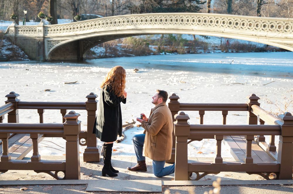 Man proposes to woman on bridge in Central Park, New York, with frozen lake and Bow Bridge in background.