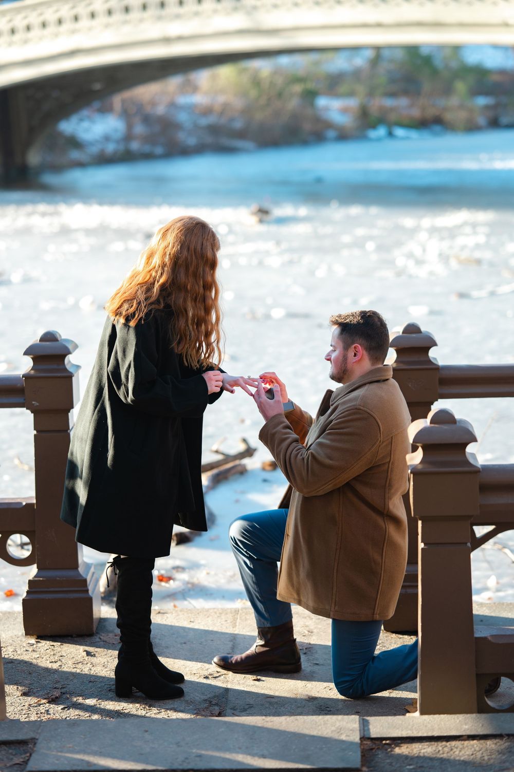 Man proposes to woman on one knee by a frozen lake with a bridge in Central Park, NYC.