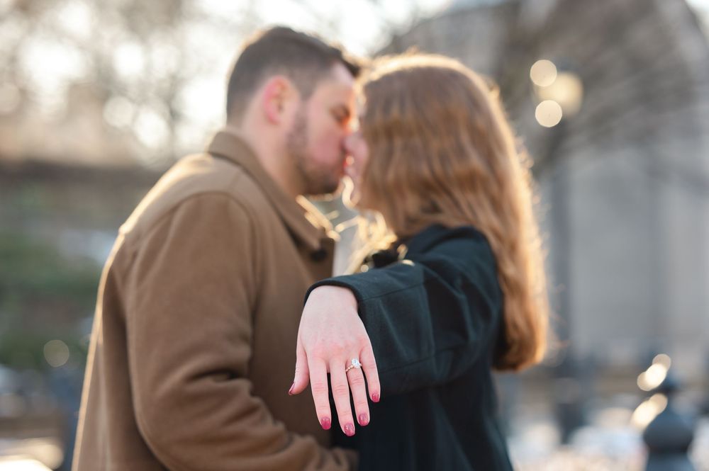 Couple kissing while showing off engagement ring outdoors in soft bokeh light.