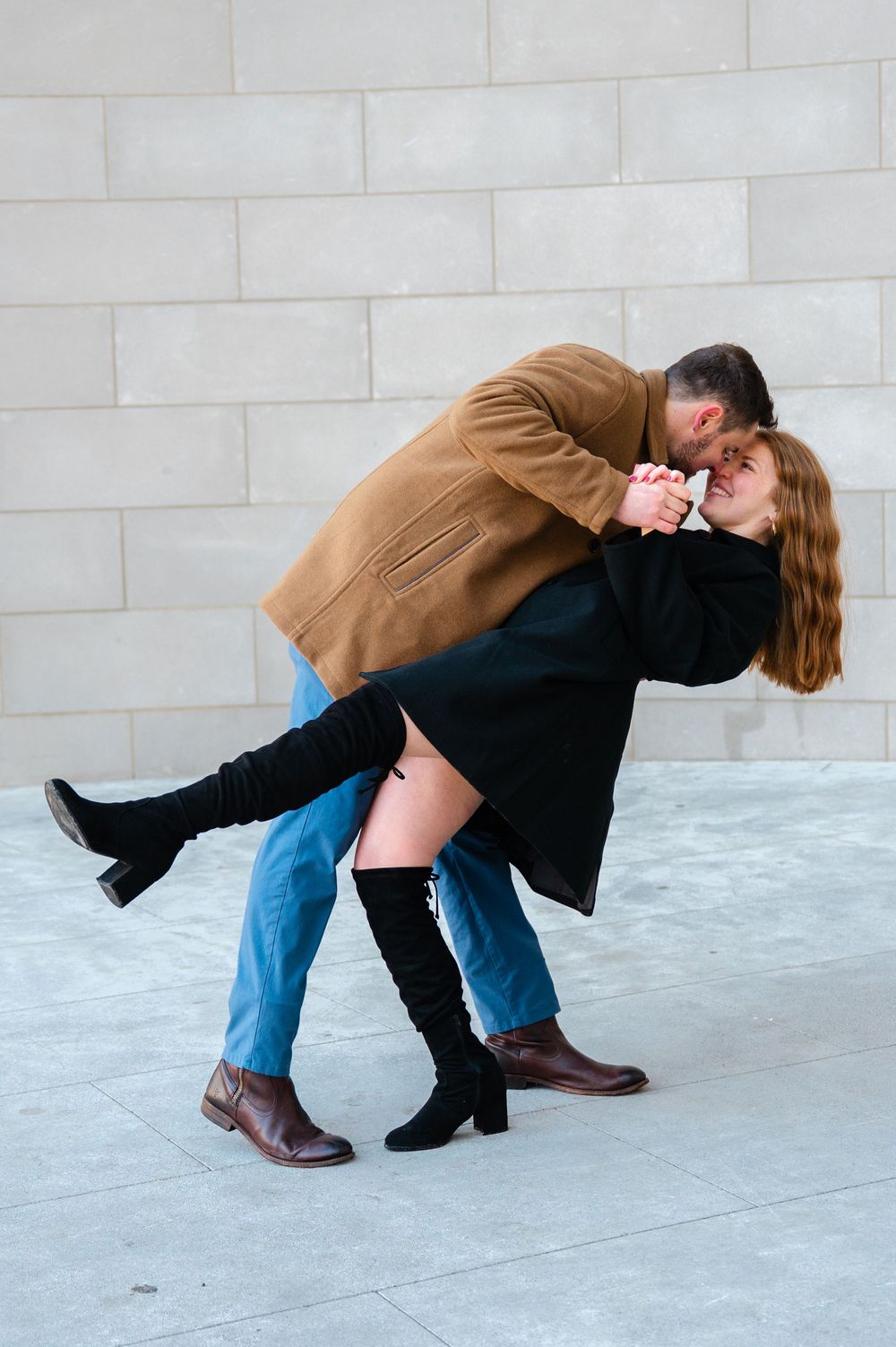 Couple dancing romantically, man dipping woman in black coat and knee-high boots against a wall.