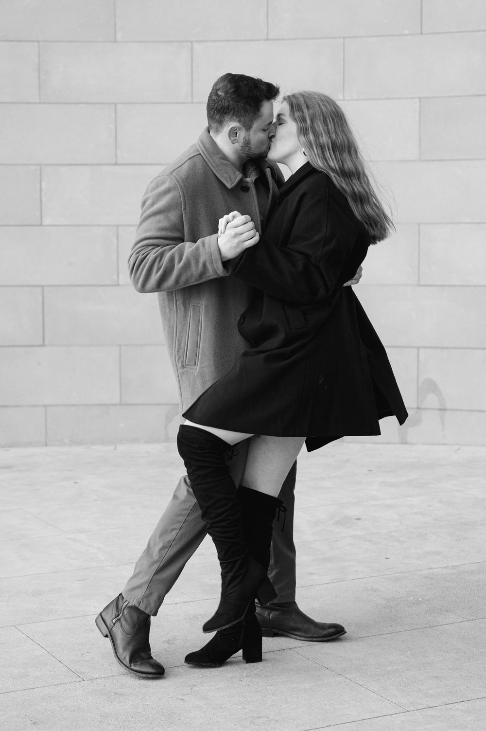 Black and white photo of a couple dancing tango and kissing, woman in thigh-high boots and black coat.