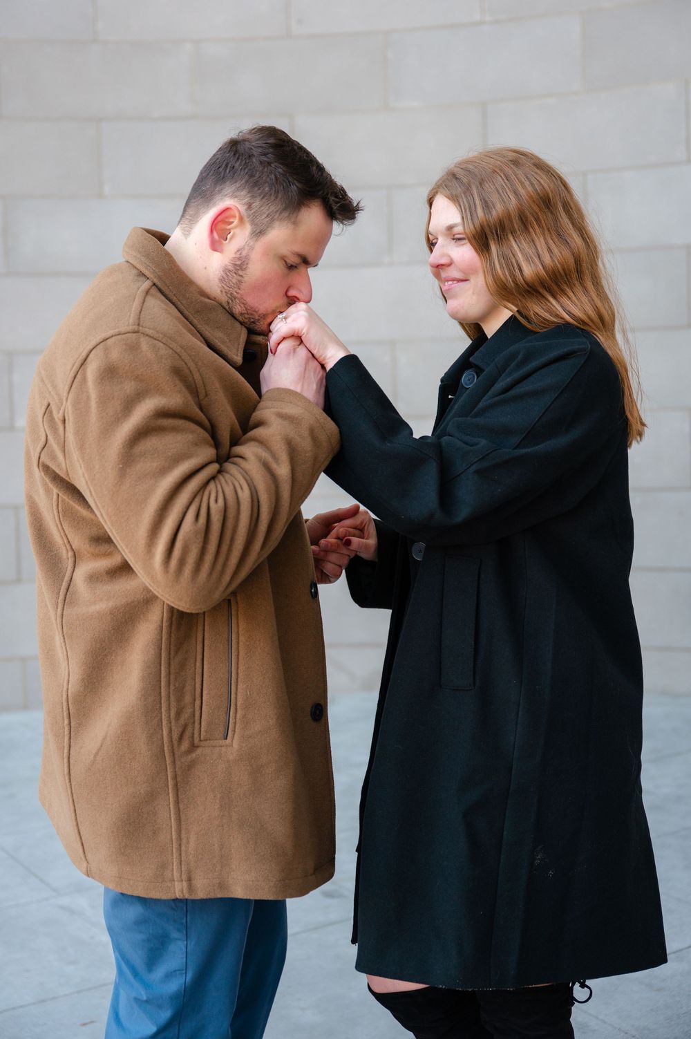 Man in tan coat kissing woman's hand during romantic engagement moment against stone wall backdrop.