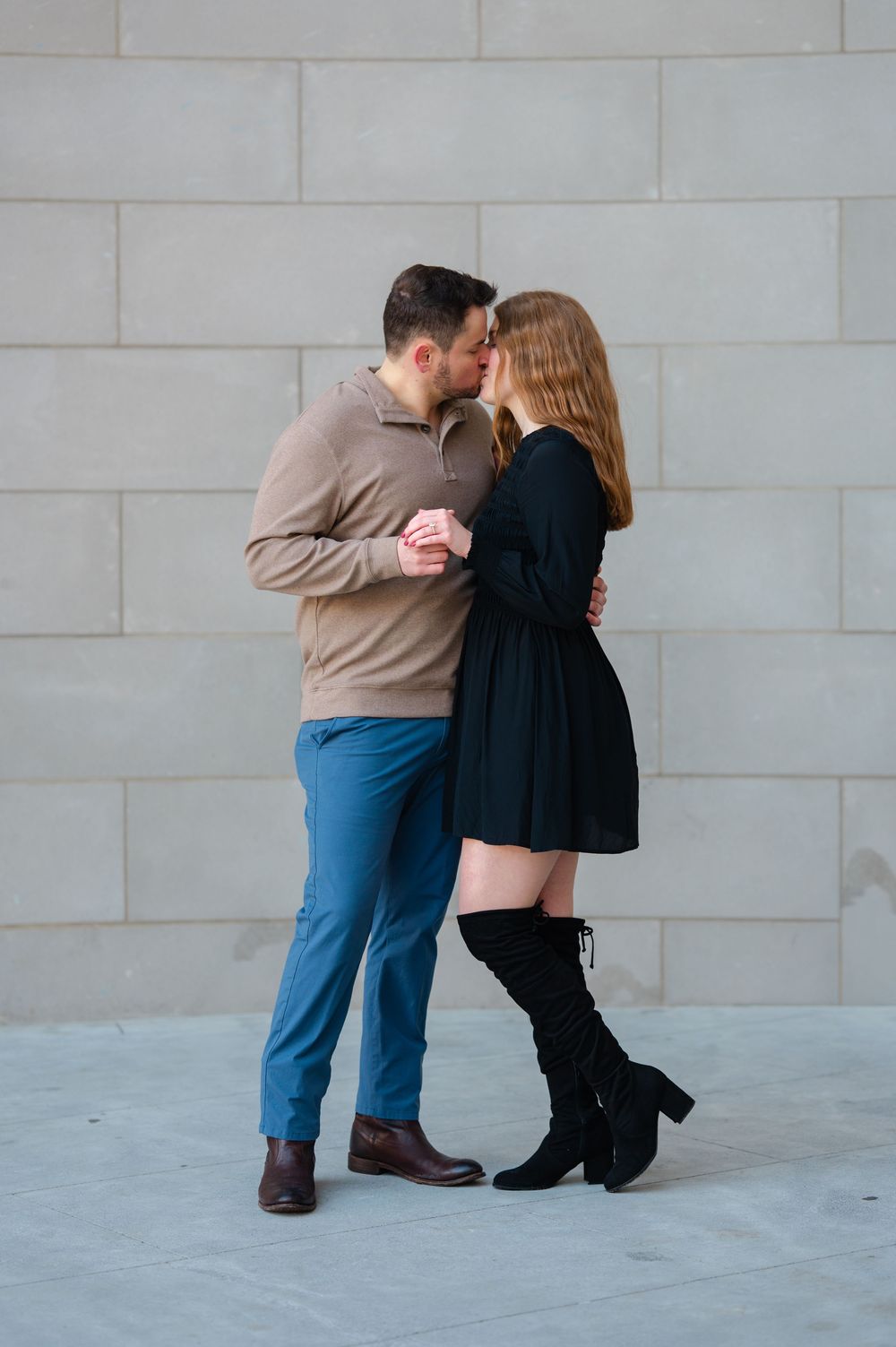 Couple sharing a romantic kiss against a gray wall, woman in black dress and knee-high boots, man in tan sweater and blue pants.