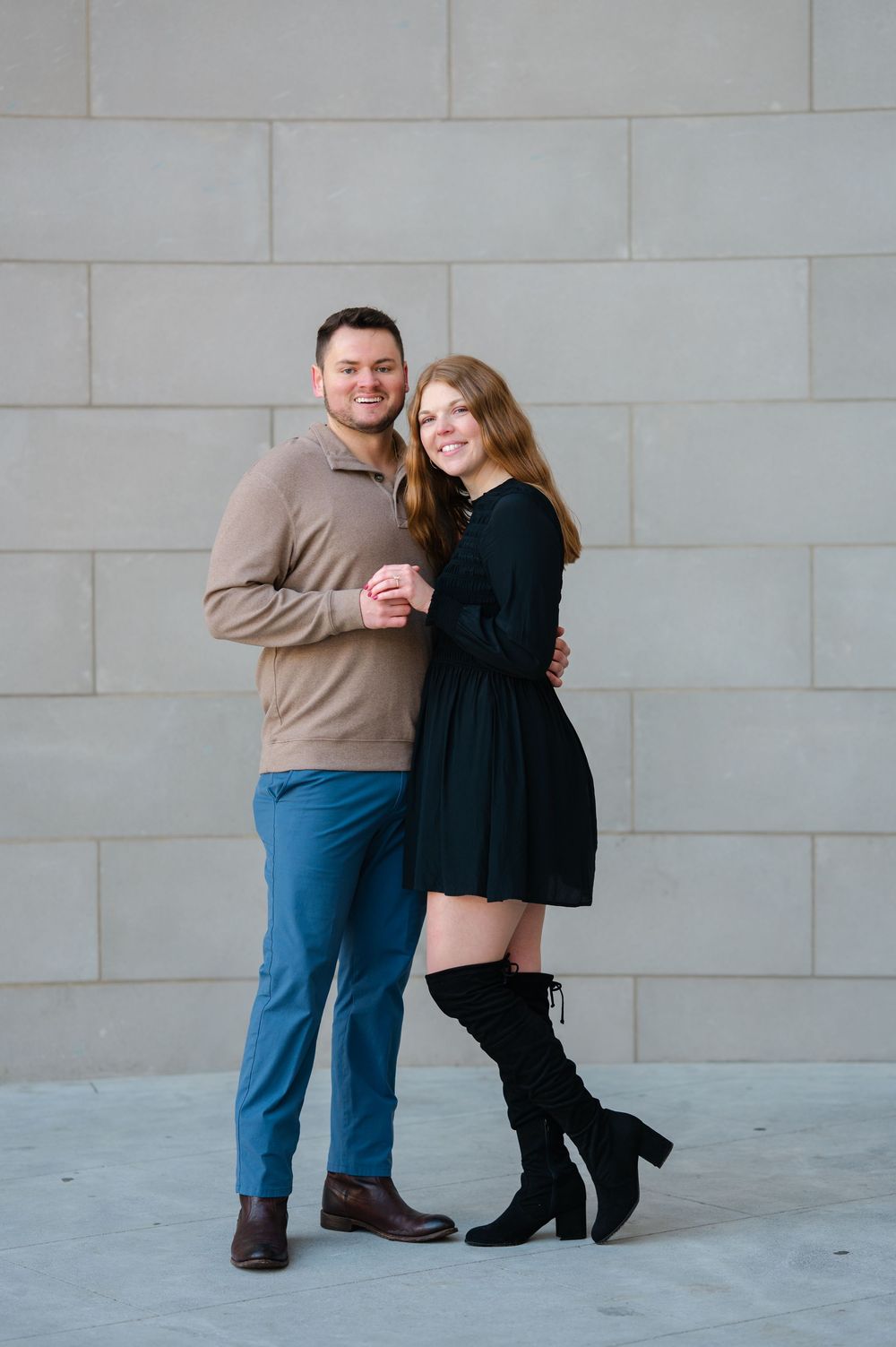 A smiling couple poses together against a gray stone wall, the woman in a black dress and boots, the man in a tan sweater and blue pants.