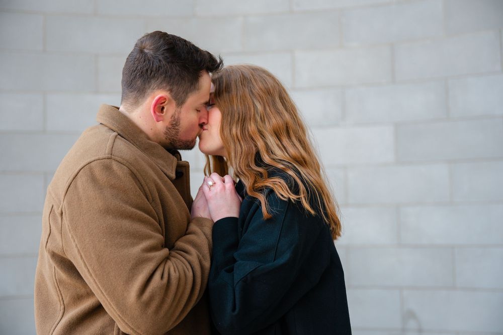 A couple sharing an intimate moment, nearly kissing, against a white brick wall in winter coats.