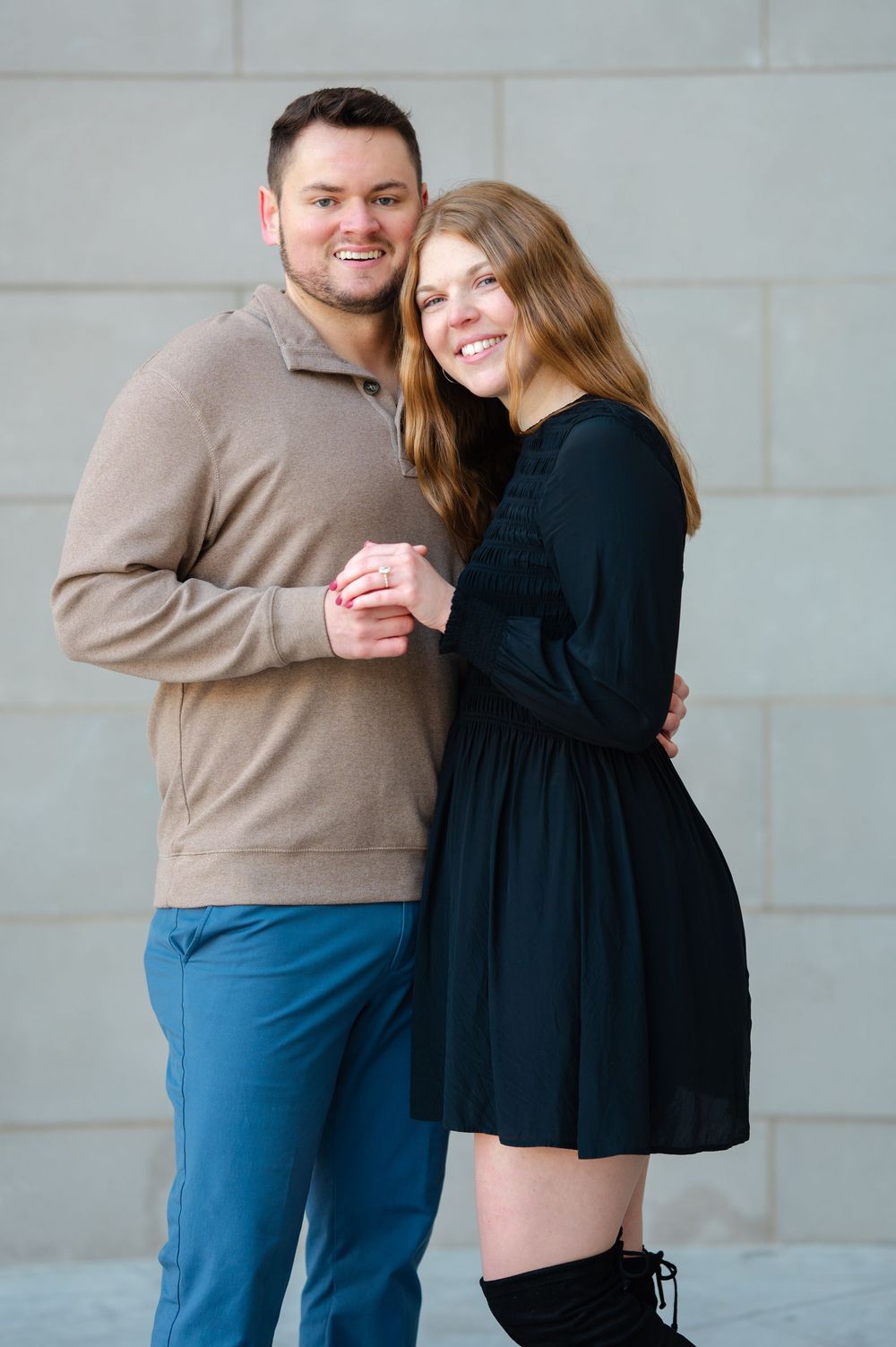 Smiling couple posing together, woman in black dress and man in tan sweater with blue pants against gray wall.