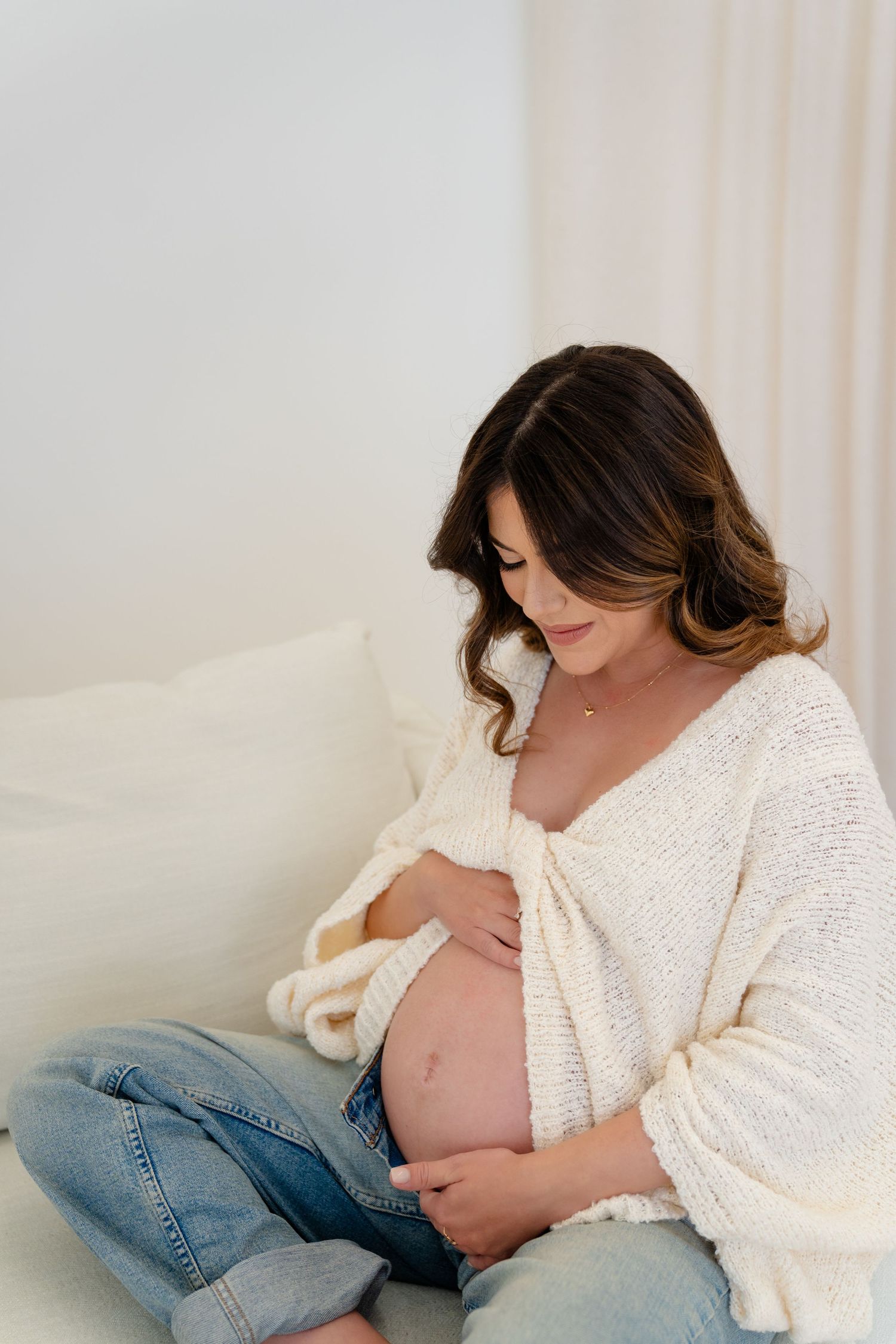 Pregnant woman in white knit sweater and jeans sitting on bed cradling her baby bump with a serene smile.
