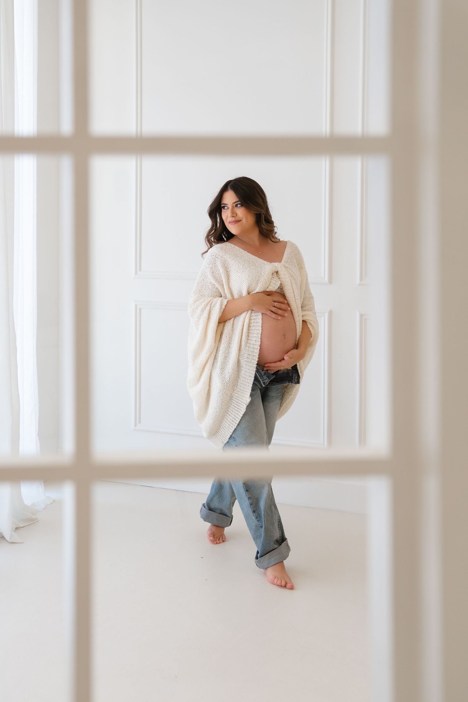 Pregnant woman in cream cardigan and jeans cradling baby bump, standing barefoot in bright white studio.