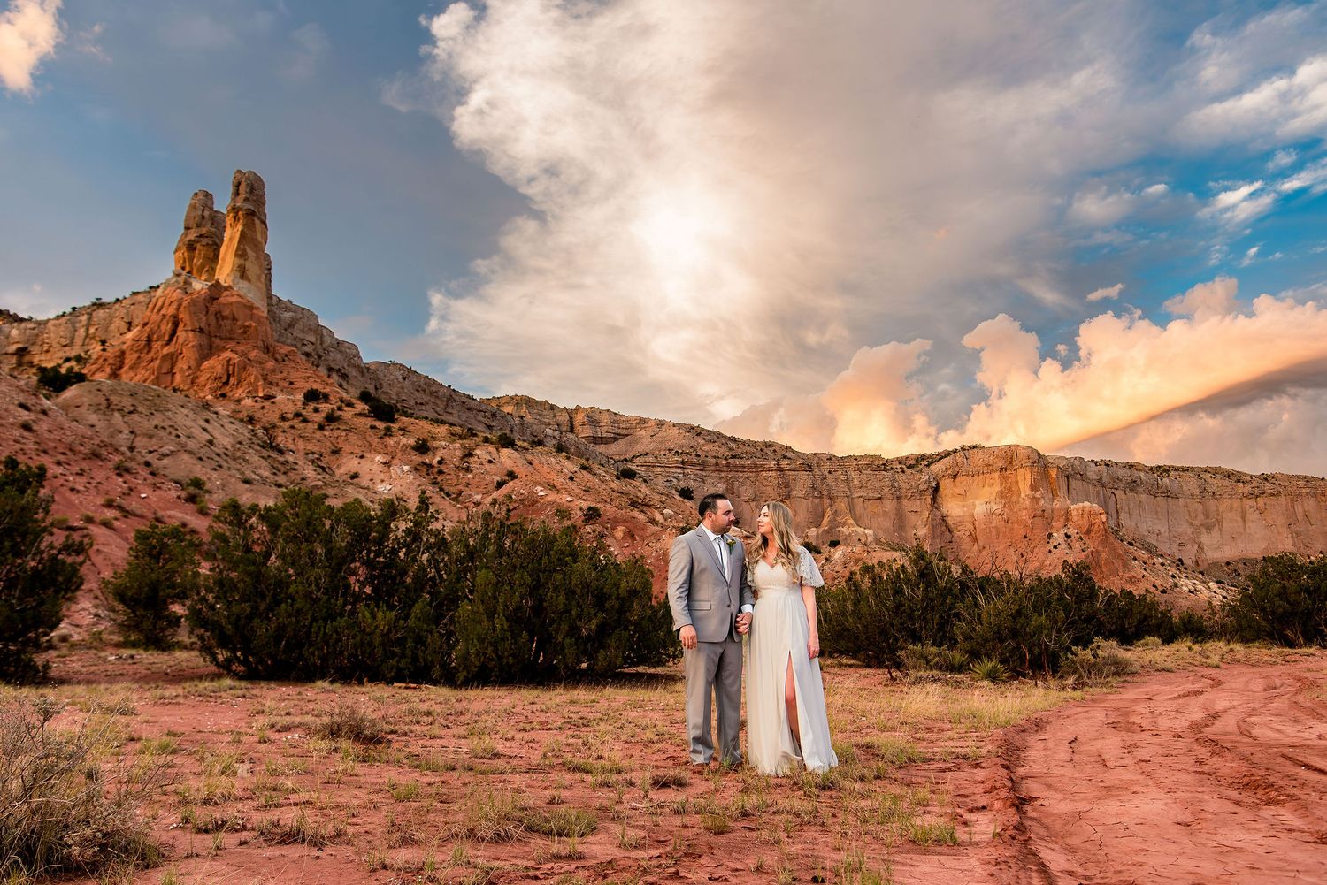 Couple in wedding attire poses before red rock formations and dramatic sunset sky in a desert landscape at Ghost Ranch in New Mexico.