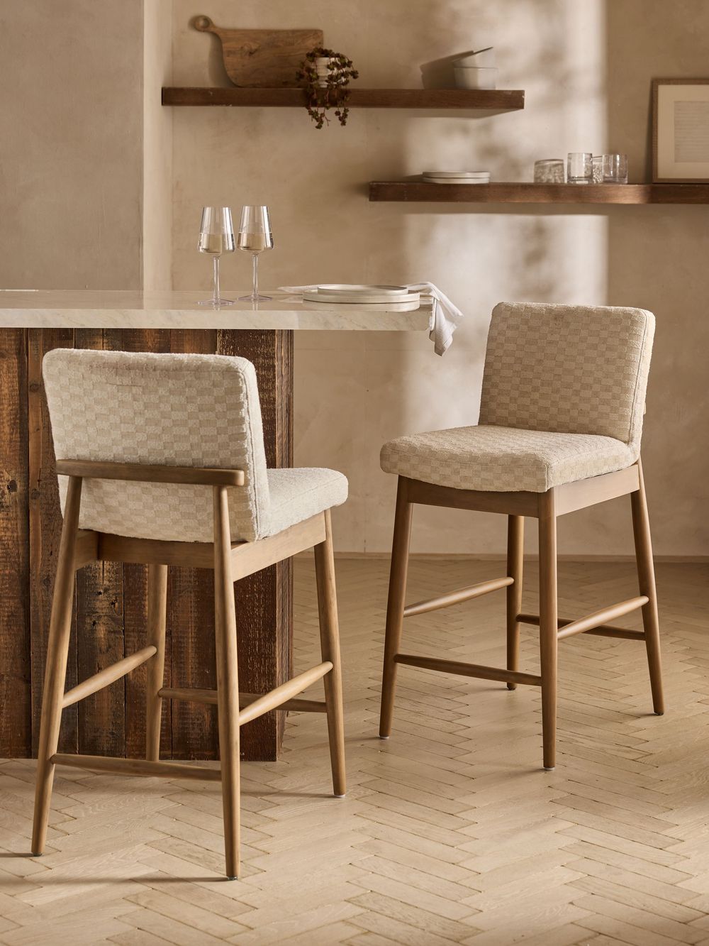 Two upholstered bar stools with wooden frames and checkered fabric beside a rustic kitchen island on herringbone floors.
