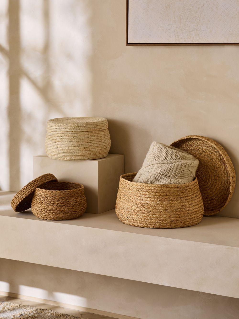 Natural woven seagrass and palm leaf baskets with lids displayed on beige shelves in a minimalist, sun-lit room.