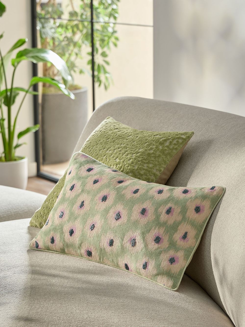 Two decorative cushions on a sofa, one green textured and one floral patterned in pink and green, with a plant in the background.