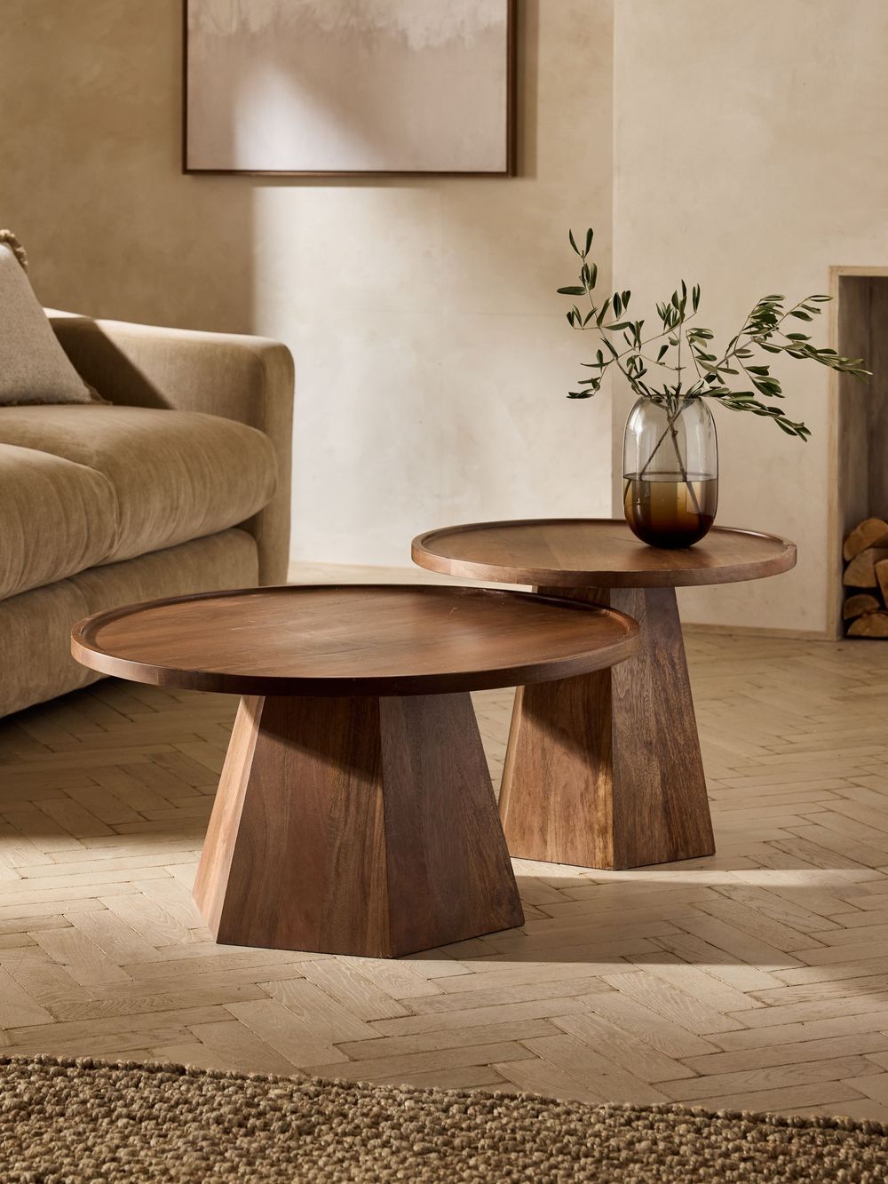 Two round dark walnut wood nesting coffee tables with geometric pedestal bases in a warm neutral living room setting.