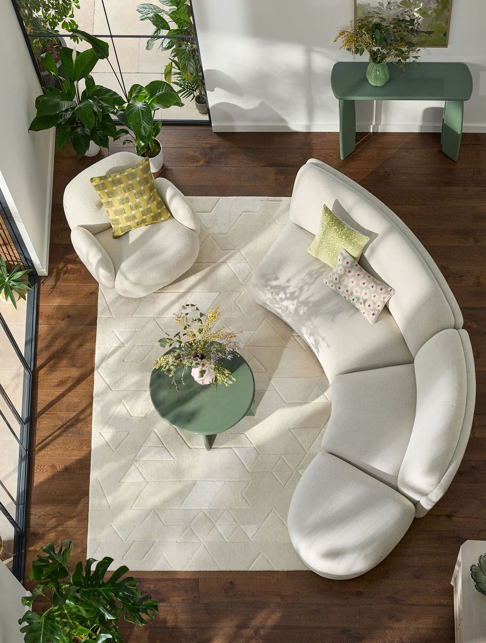 Aerial view of a curved white sectional sofa with green accents, round coffee table, and lush plants on hardwood floors.