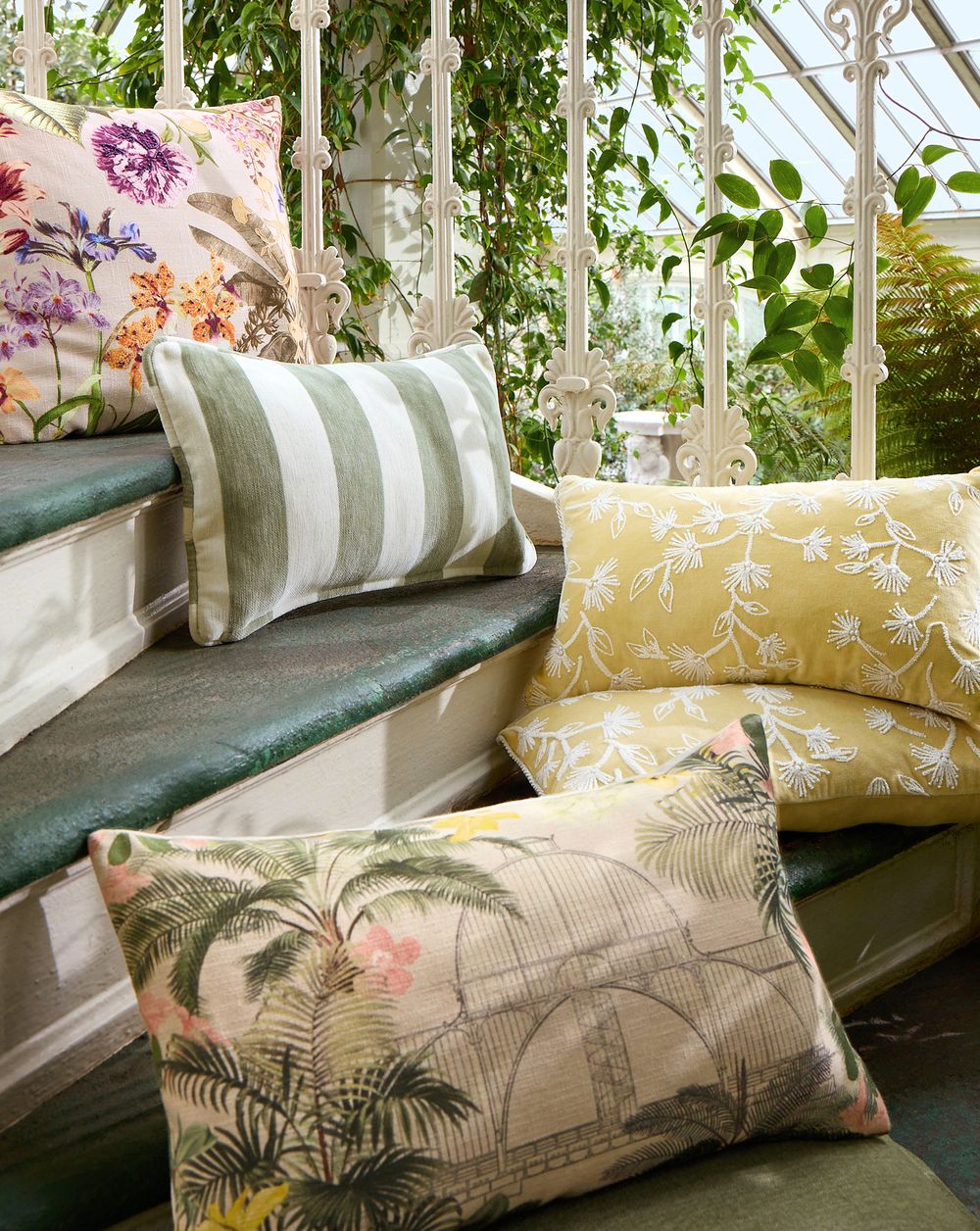 Decorative botanical print cushions in floral, stripe, and tropical palm designs arranged on a bench in a sunlit greenhouse.