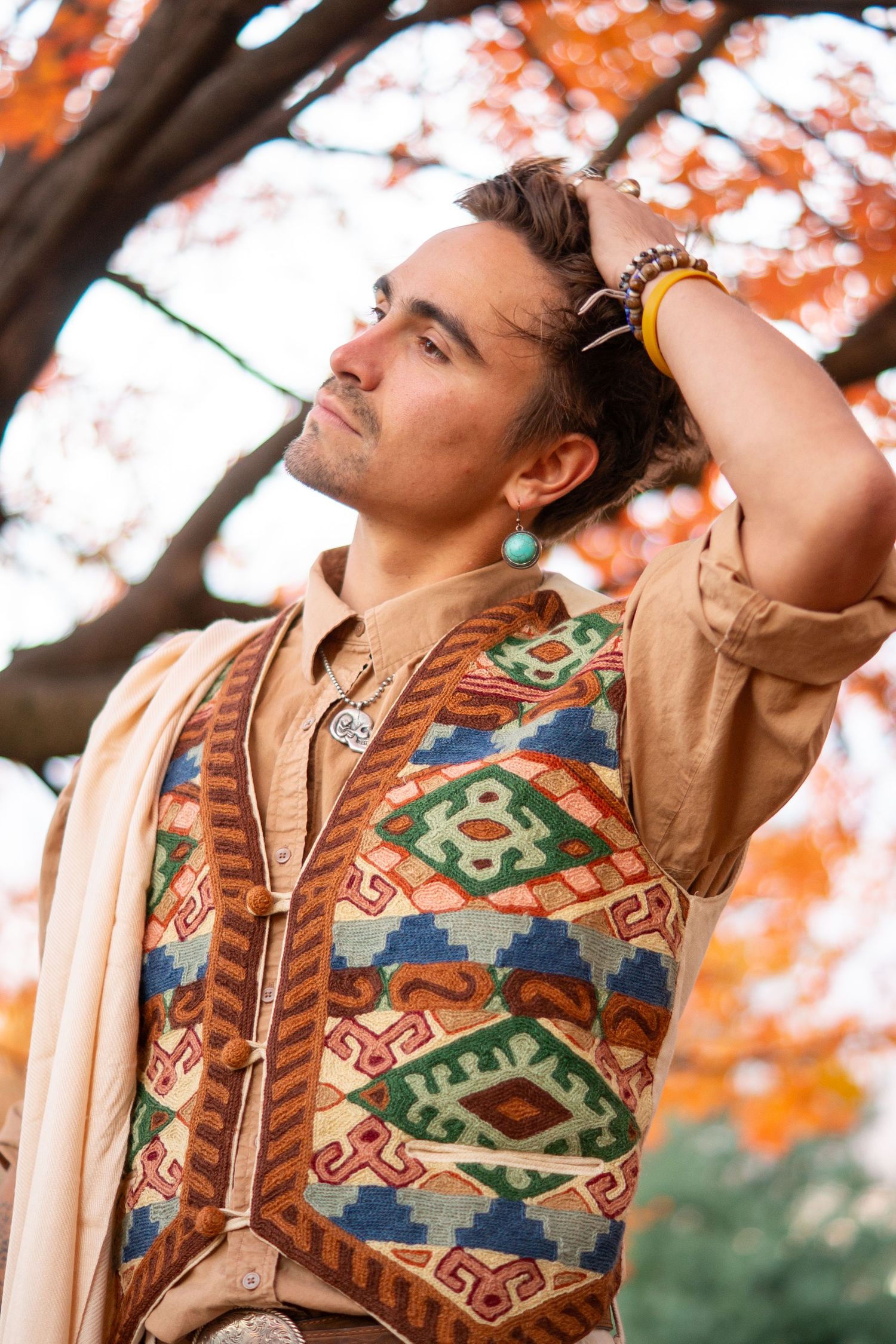 Man in colorful geometric patterned vest and beaded bracelets poses outdoors among autumn foliage trees.