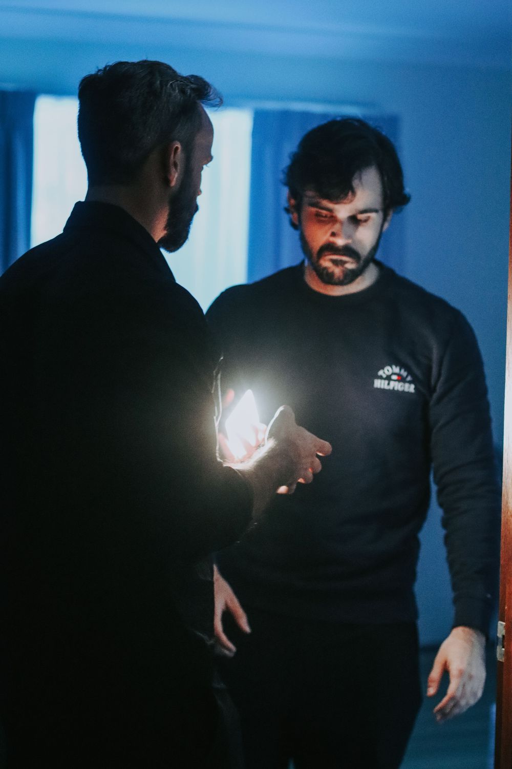 Two bearded men in dark clothing examine a glowing light source in a dimly lit blue room.