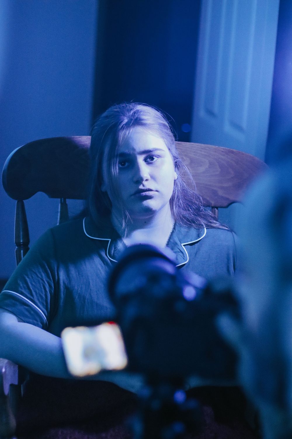 Young woman with long blonde hair sits in wooden chair under blue lighting, filmed by camera in moody room.