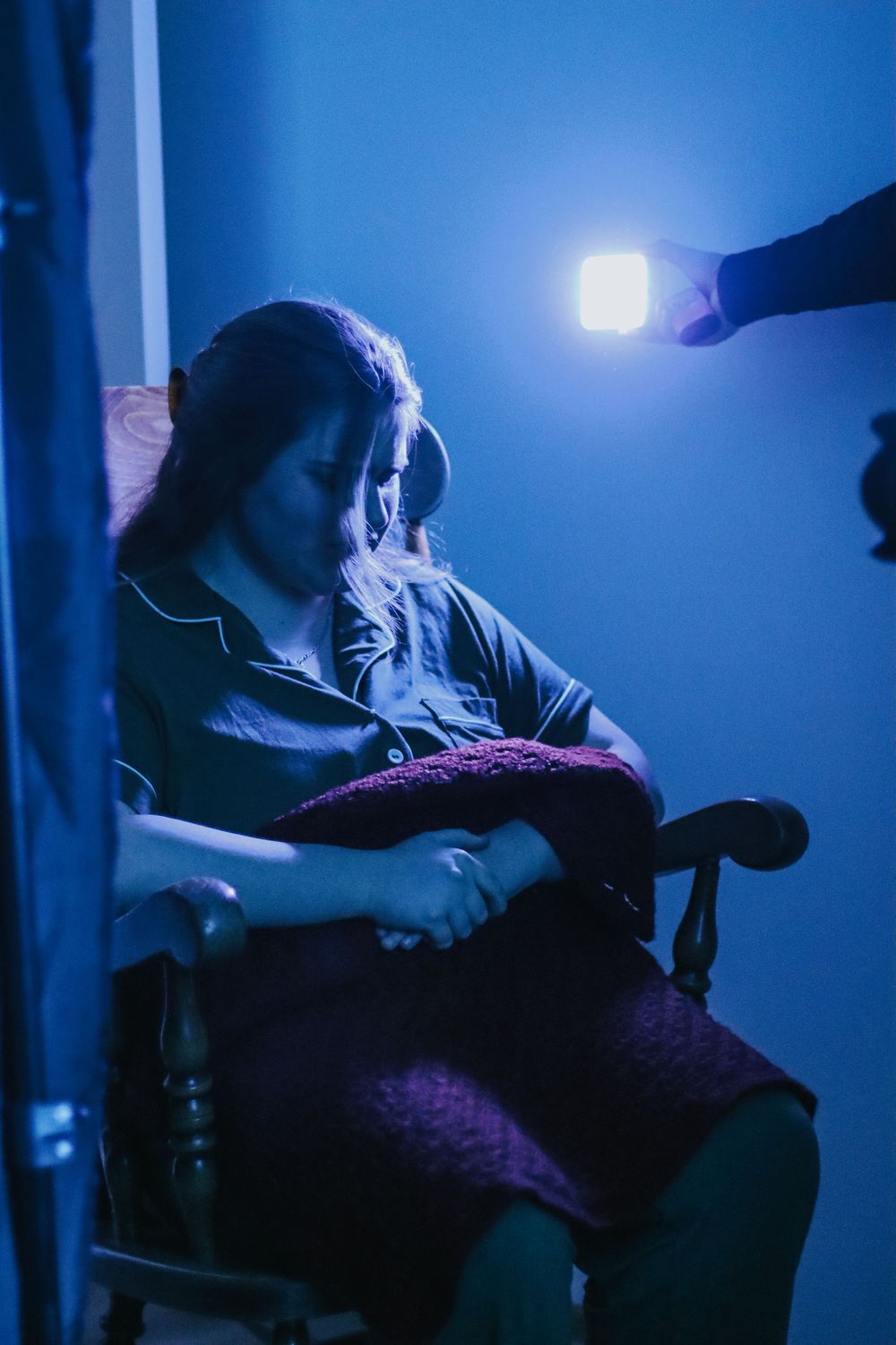 Woman sitting in rocking chair holding pillow in dark blue room illuminated by handheld light.