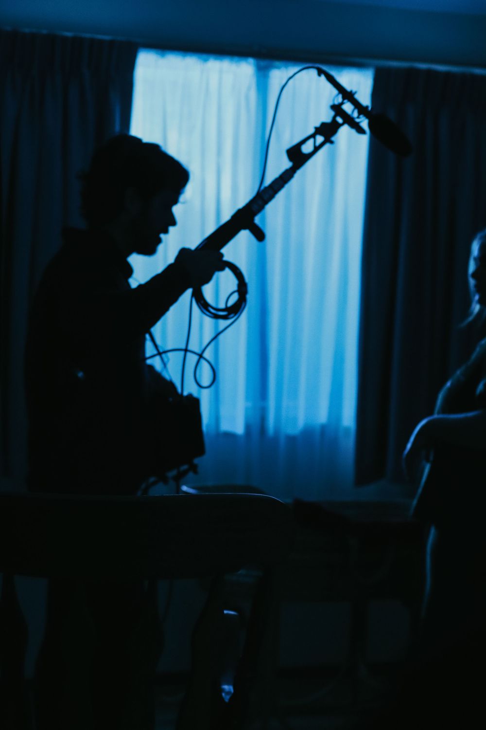 Silhouette of a guitarist playing electric guitar in a dark blue-lit room with curtains in the background.