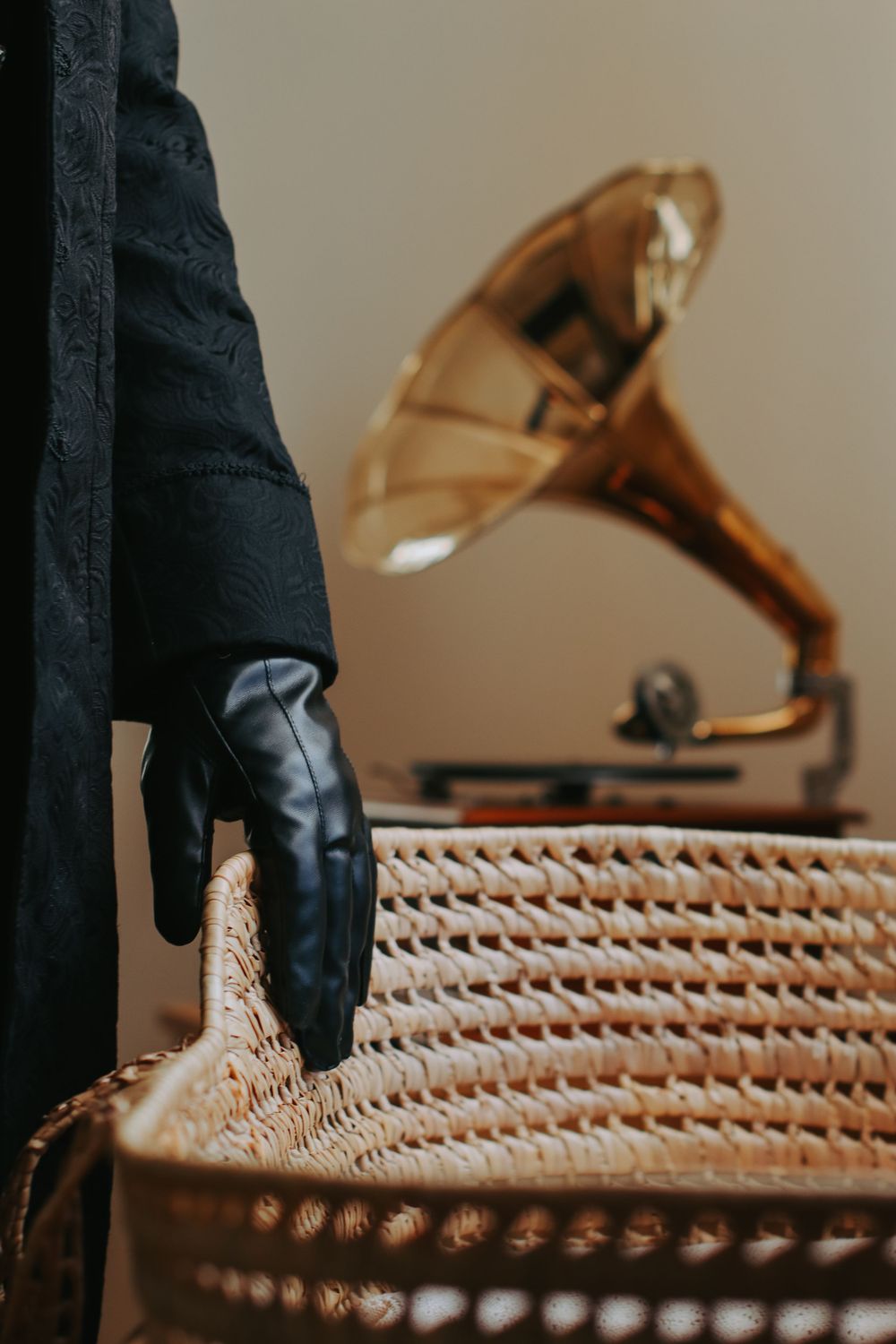 A gloved hand in black leather rests on a wicker chair beside a vintage gold gramophone in an elegant setting.