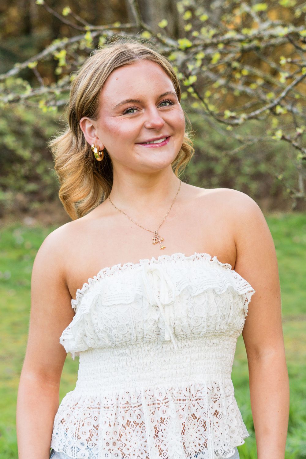 Young woman in white lace strapless dress smiling outdoors with green trees in background.
