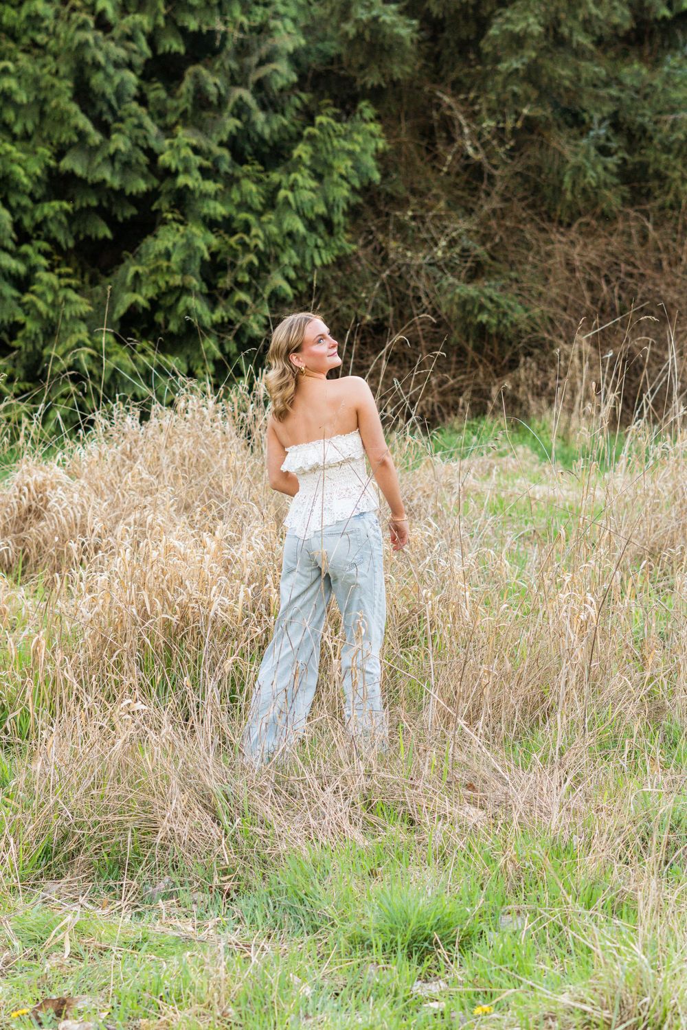 Young woman in white strapless top and striped wide-leg pants standing in tall grass field near evergreen trees.