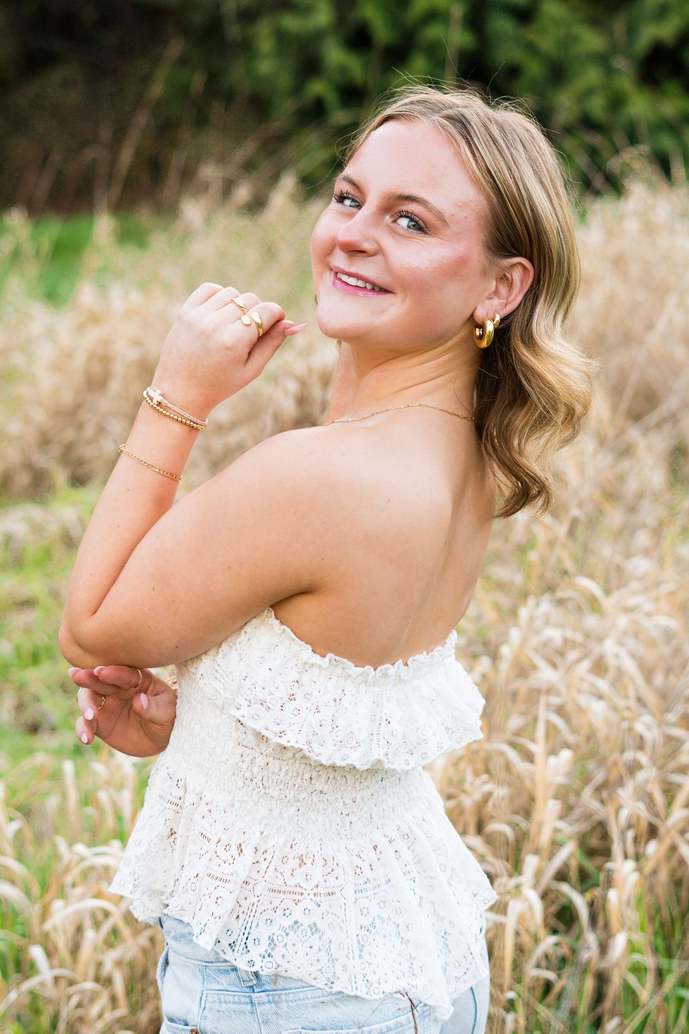 Young woman in white lace strapless top and jeans smiling over shoulder in golden field.