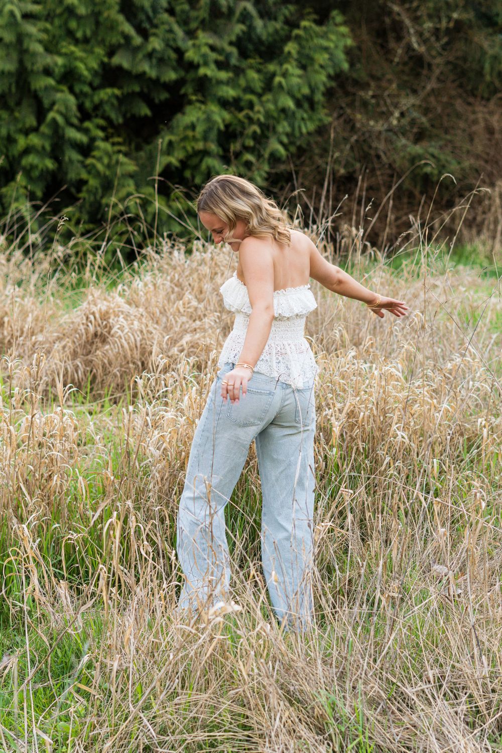 Blonde woman in white ruffled top and light jeans standing in tall golden grass field.