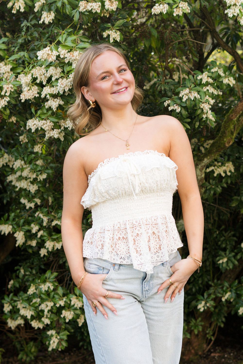 Young blonde woman smiling in white lace strapless top and jeans posed before flowering bushes.