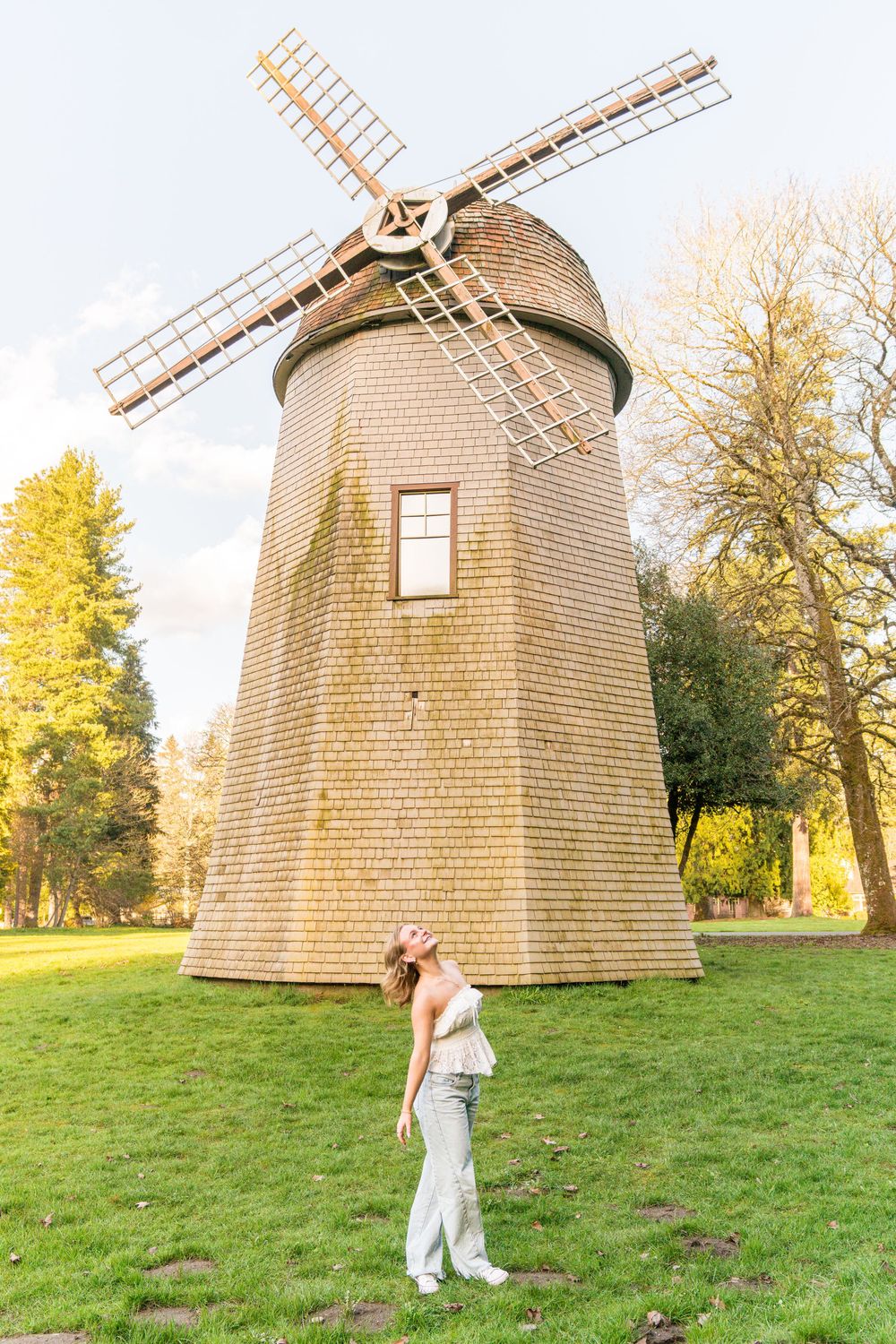 Smiling blonde woman posing beside a tall historic windmill in a green park at golden hour.