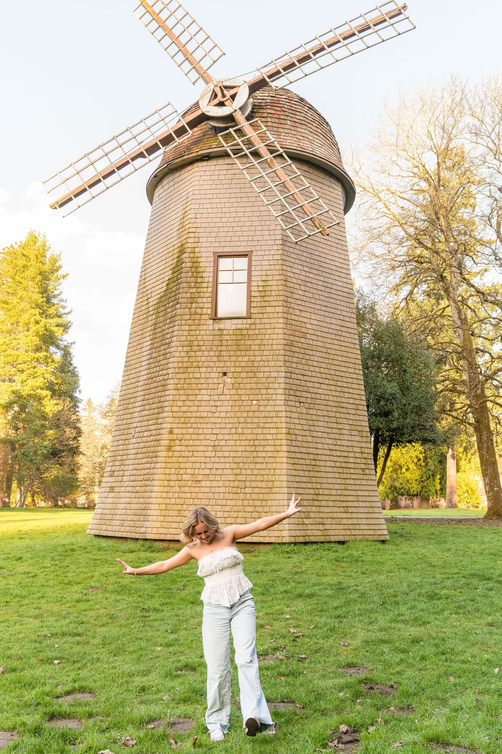 Young woman posing with arms outstretched in front of a historic wooden windmill surrounded by autumn trees.