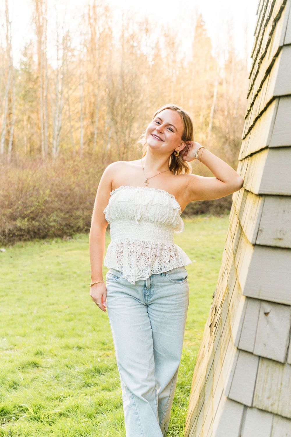 Smiling young woman in white lace tube top and light jeans leaning against a wooden building outdoors in autumn.