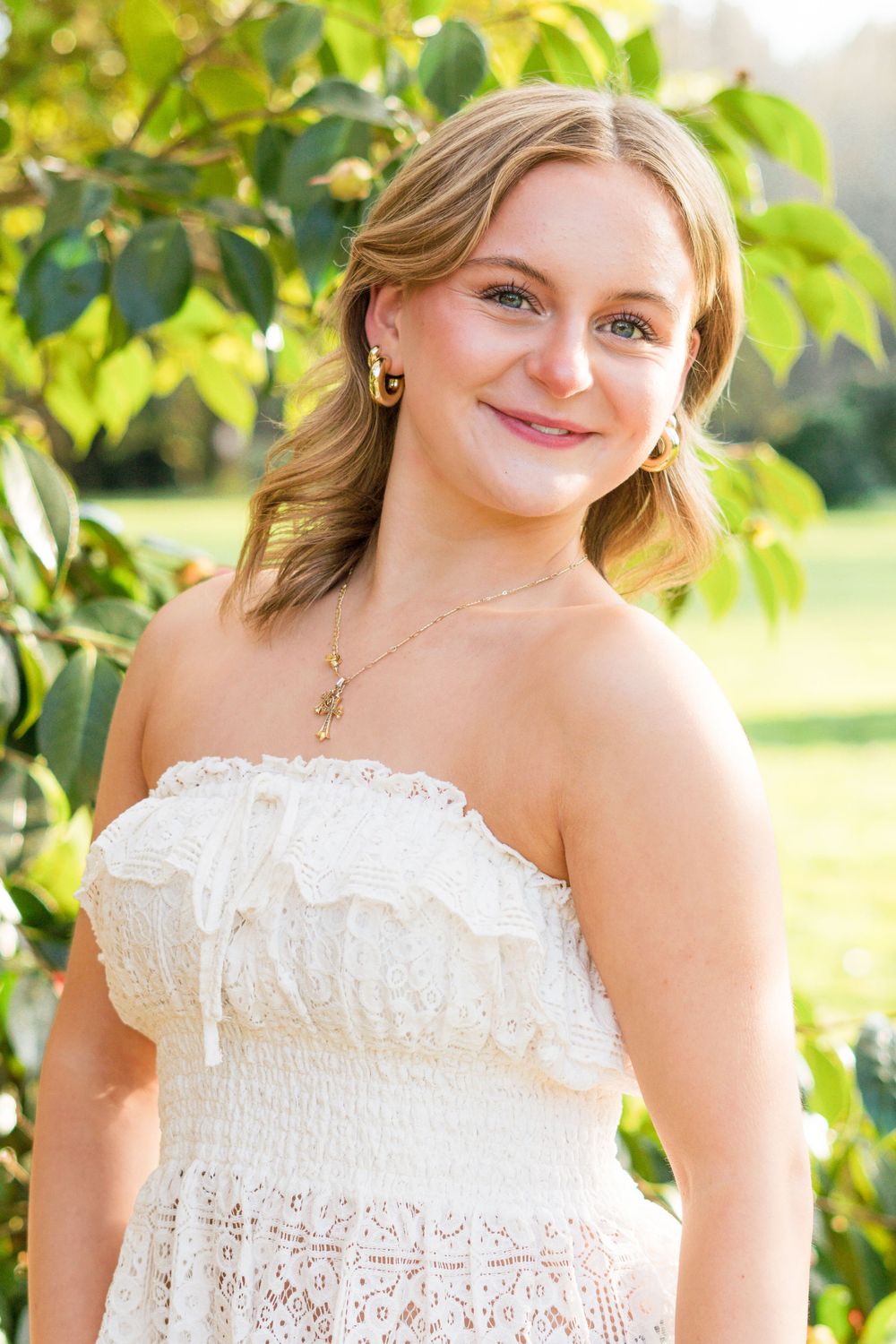 Young blonde woman in white strapless lace dress smiling outdoors among green leaves on a sunny day.