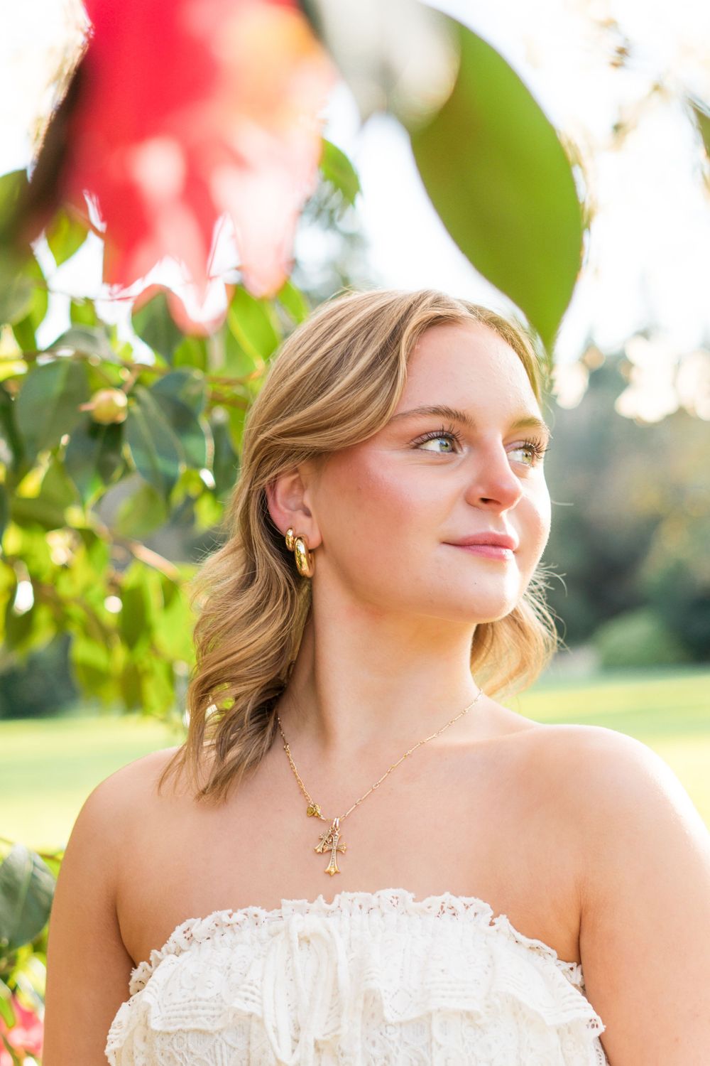 Young blonde woman in white strapless dress wearing gold jewelry, gazing upward near red flowers outdoors.