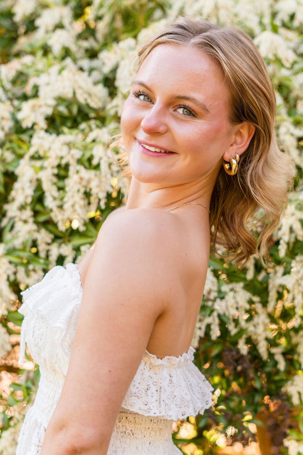 Smiling blonde woman in white strapless lace dress with gold hoop earrings posed before blooming white flowers.