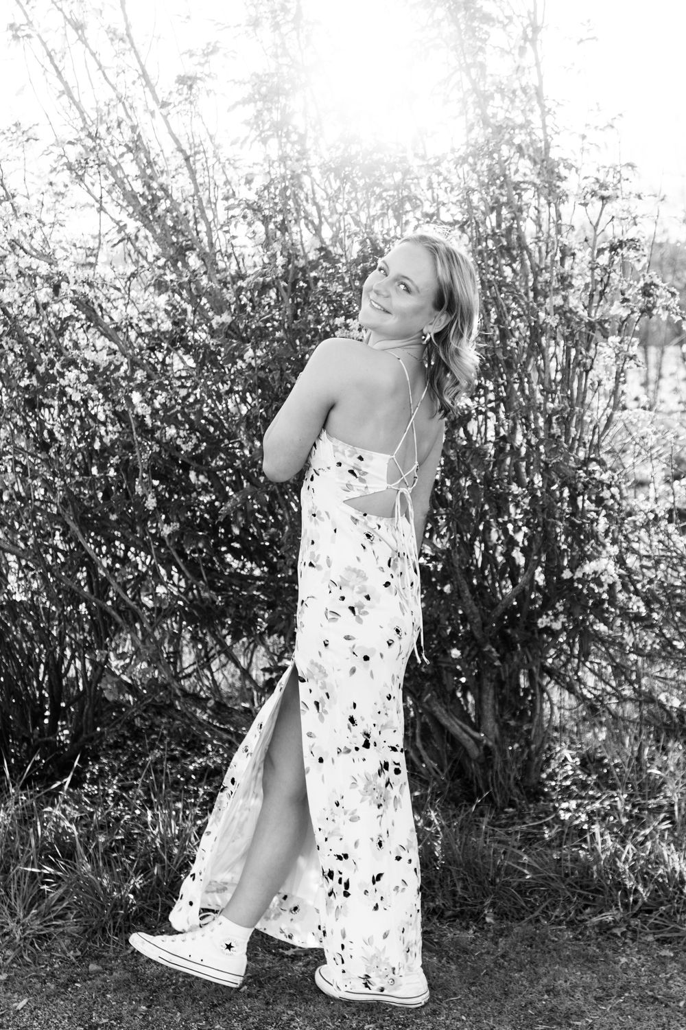 Blonde woman in floral maxi dress and sneakers posing outdoors near bushes in black and white photo.