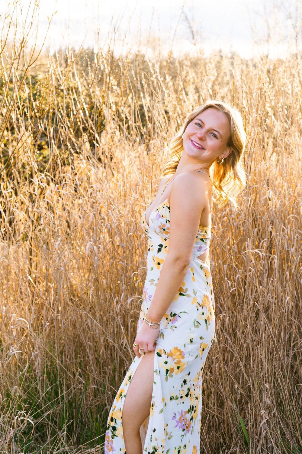 Smiling blonde woman in floral maxi dress standing in golden tall grass field at sunset.