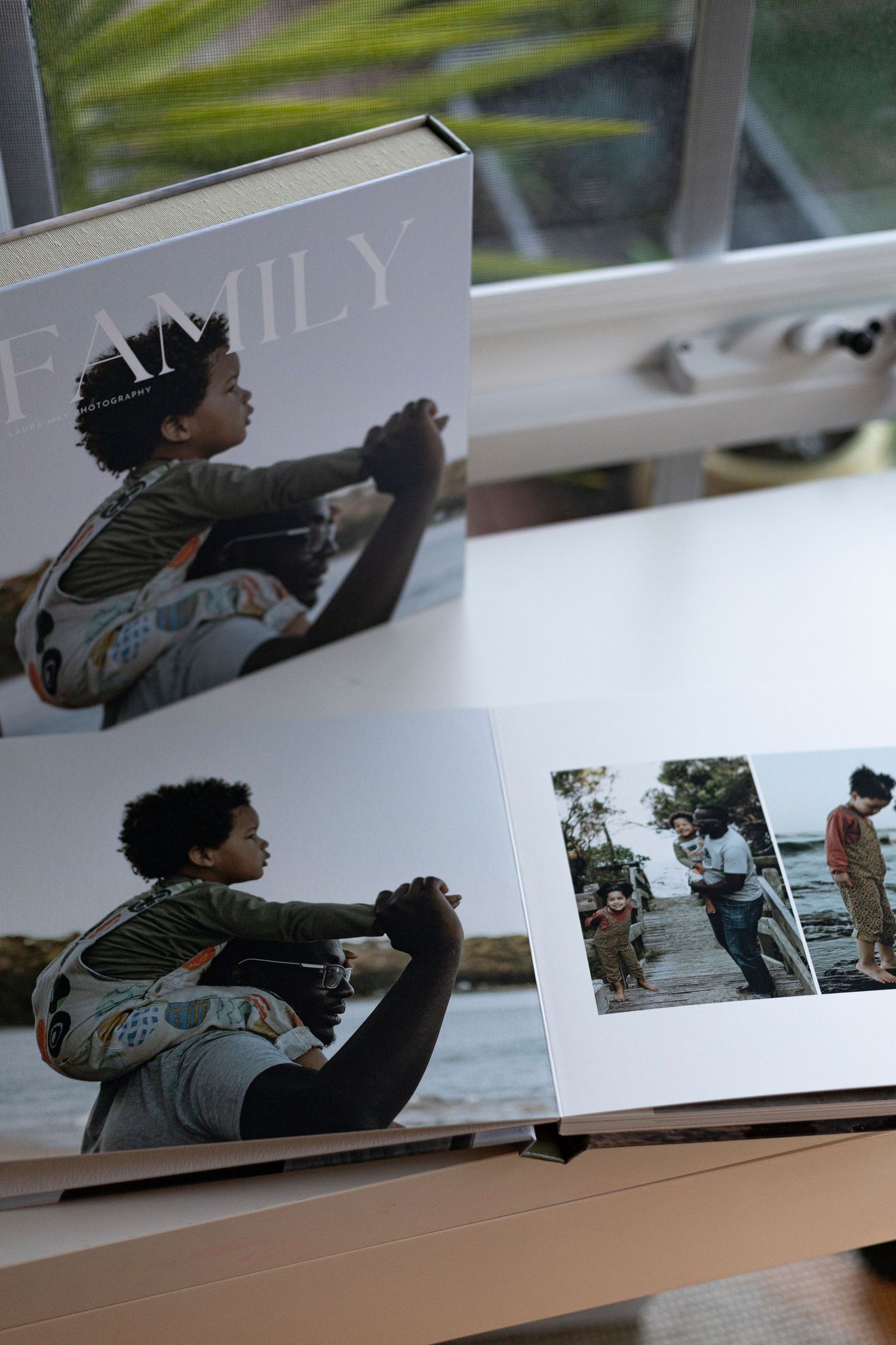 Family photo book open on a wooden table showing a child riding on an adult's shoulders near water, with a second book behind it.