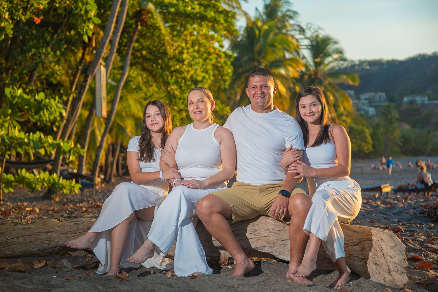Family of four smiling together on a tropical beach at sunset, dressed in white and khaki outfits.