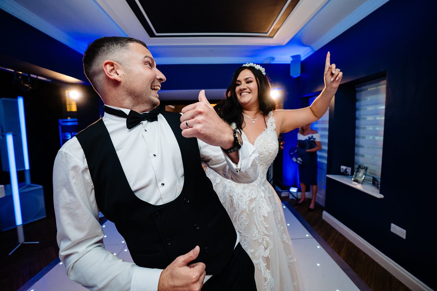 Bride and groom share a joyful first dance, laughing and pointing upward in an elegant blue-lit wedding venue.