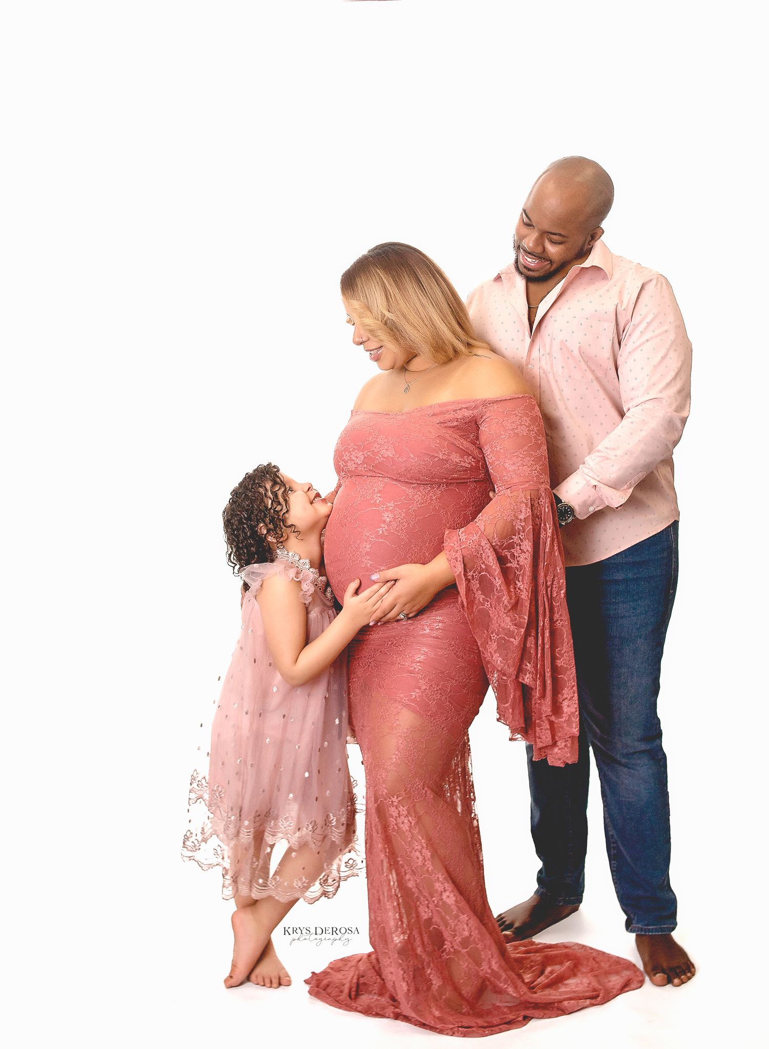 Pregnant woman in pink lace gown poses with man and young girl for maternity photo shoot on white background.