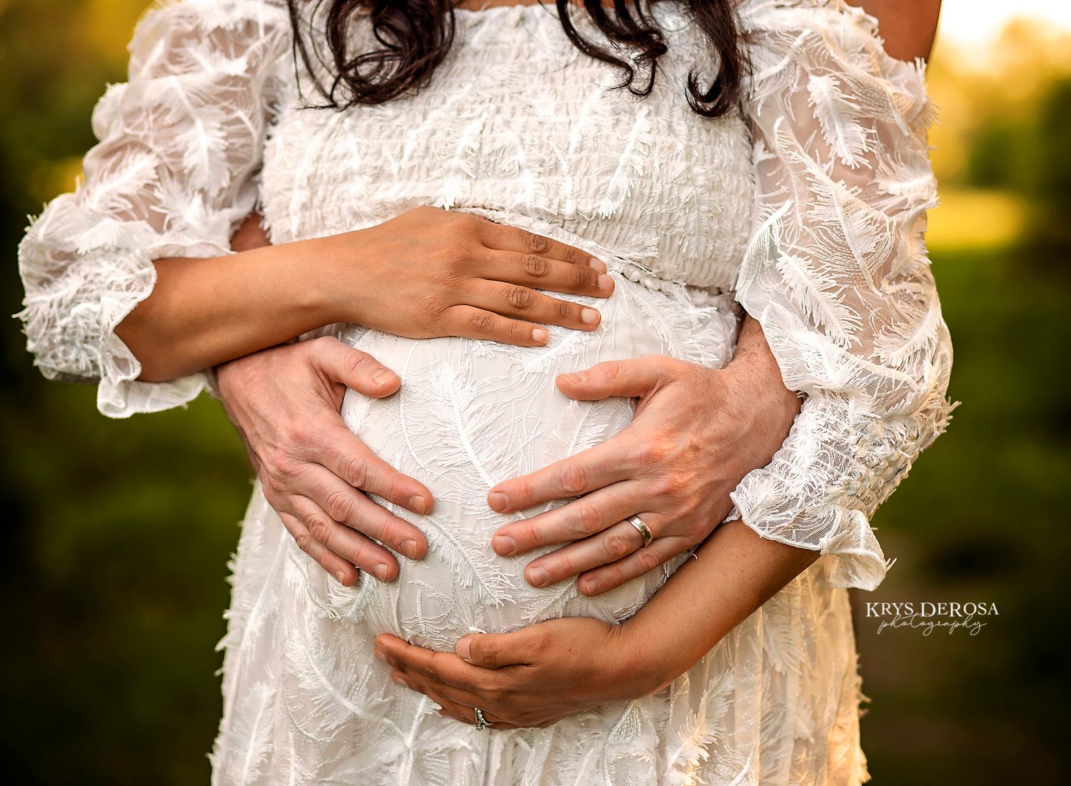 Couple cradling pregnant belly in maternity photo, woman in white lace dress, golden hour outdoor portrait.