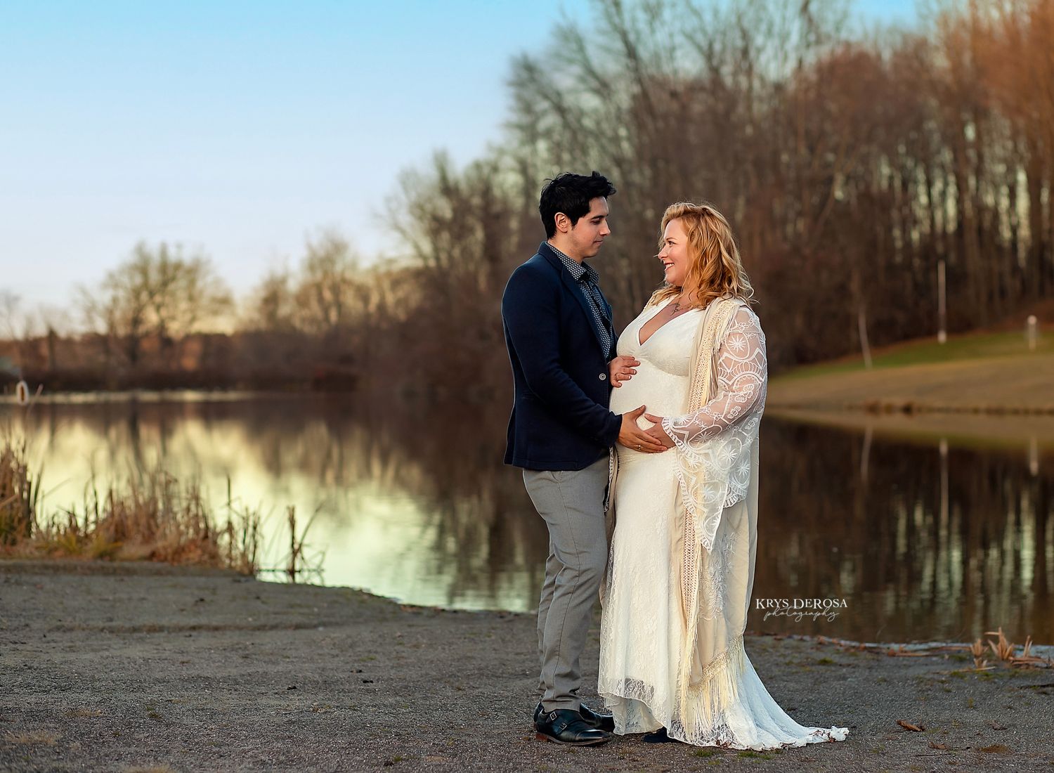 Couple posing by a lake during golden hour, woman in white lace maternity gown, man in navy blazer.