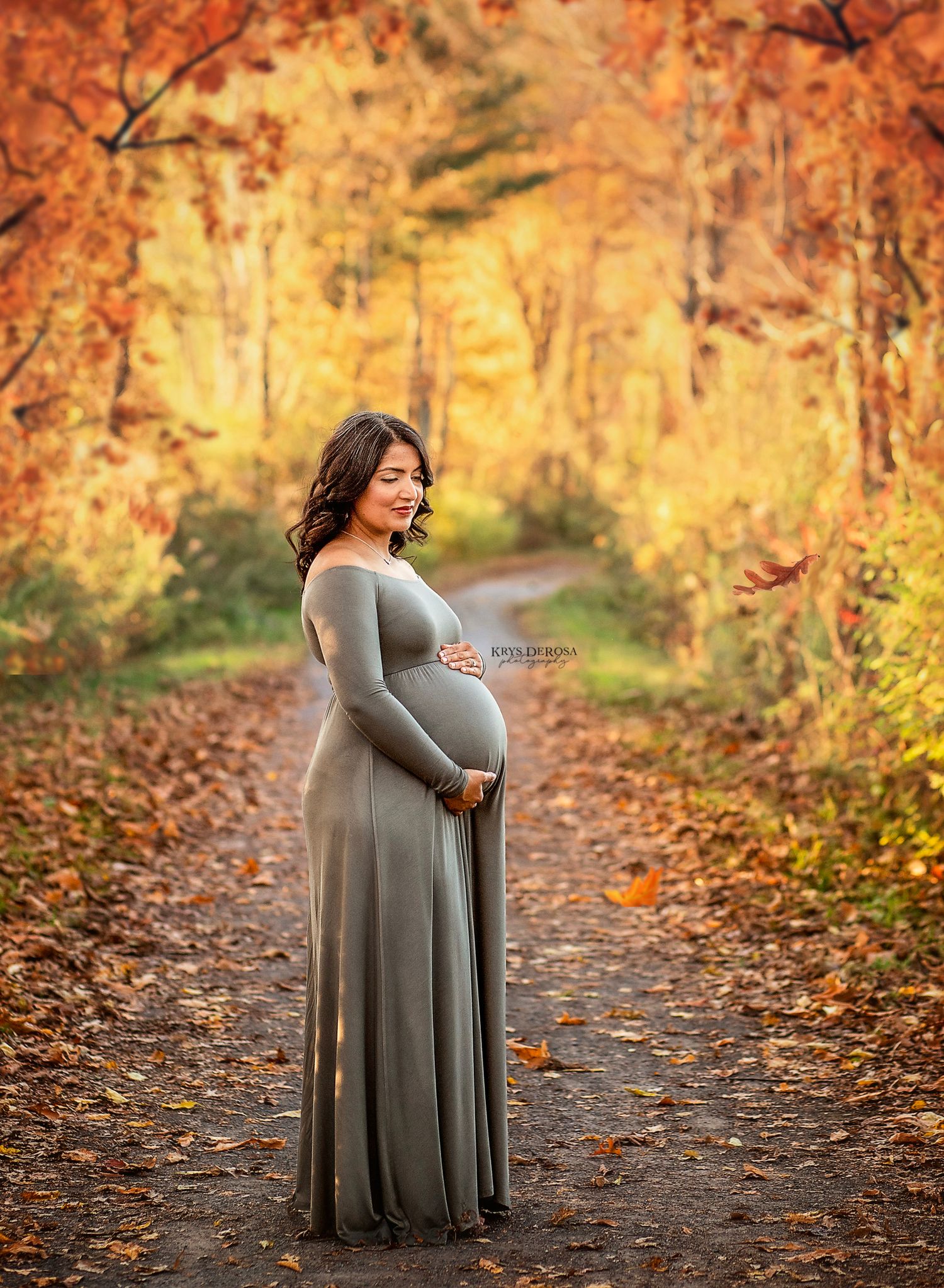 Pregnant woman in sage green gown poses on autumn forest path surrounded by vibrant orange and yellow foliage.
