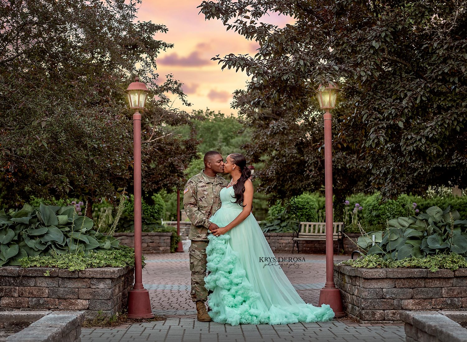 Couple sharing a romantic kiss in a garden, man in military uniform, woman in mint green gown, at sunset.