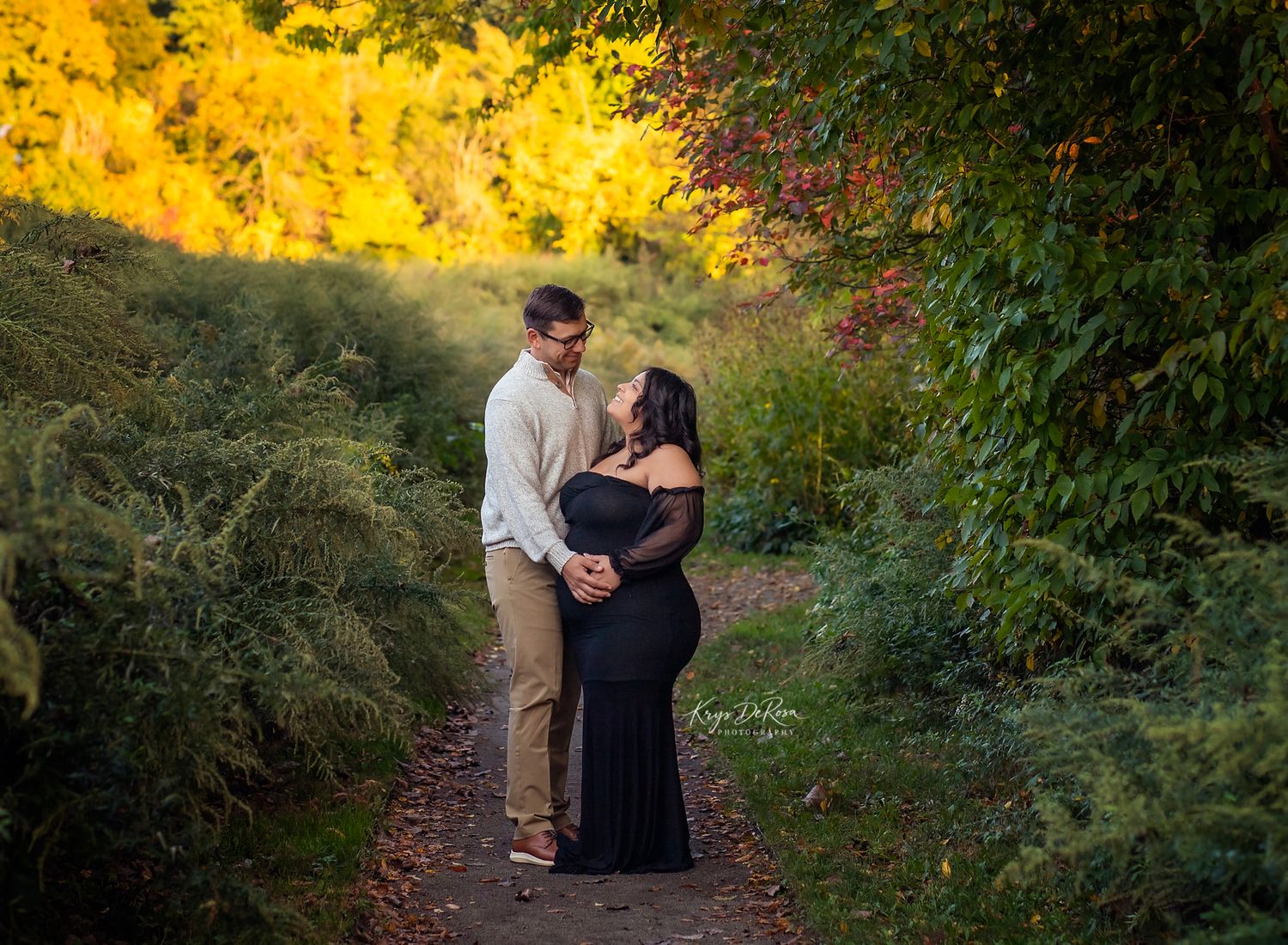 Couple sharing a romantic moment on a garden path surrounded by vibrant autumn foliage during a maternity photoshoot.