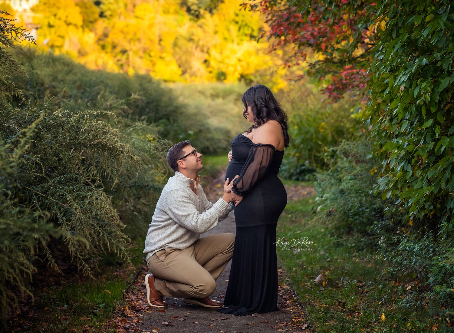 Man kneeling before pregnant woman in black gown on a scenic autumn path during an outdoor maternity photoshoot.
