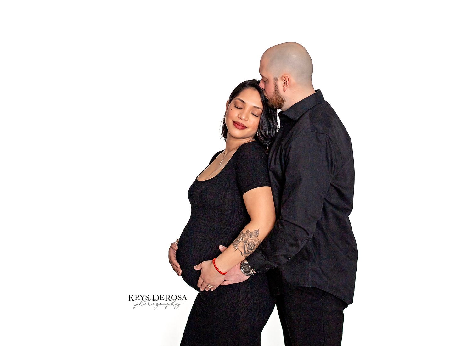 Pregnant couple in matching black outfits posing together on white studio background.